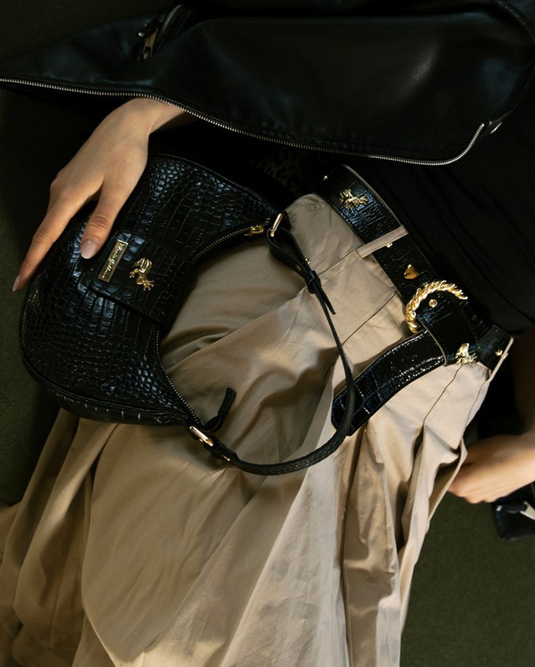 Detalle de bolso de croco negro sobre un pantalón beige.