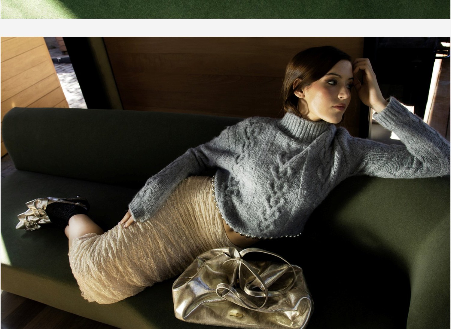 Modelo recostada en un sofá verde con sweater gris y zapatos con aplicación.