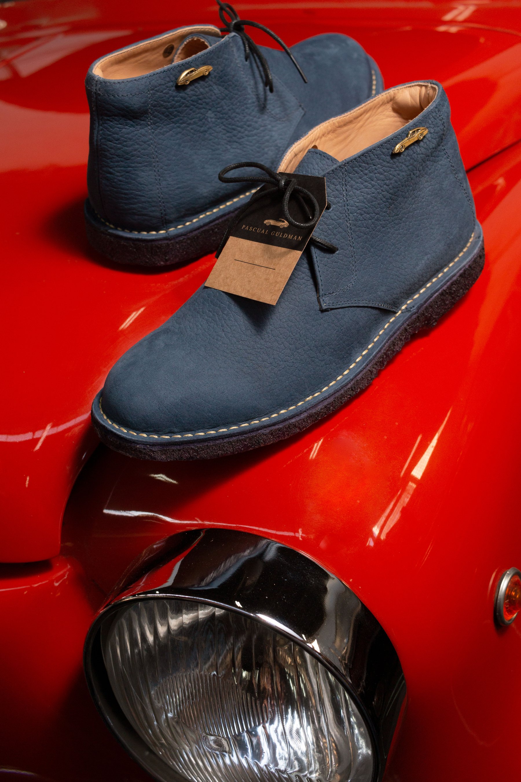 Par de botas desert de ante azul sobre el capó rojo de un auto clásico.